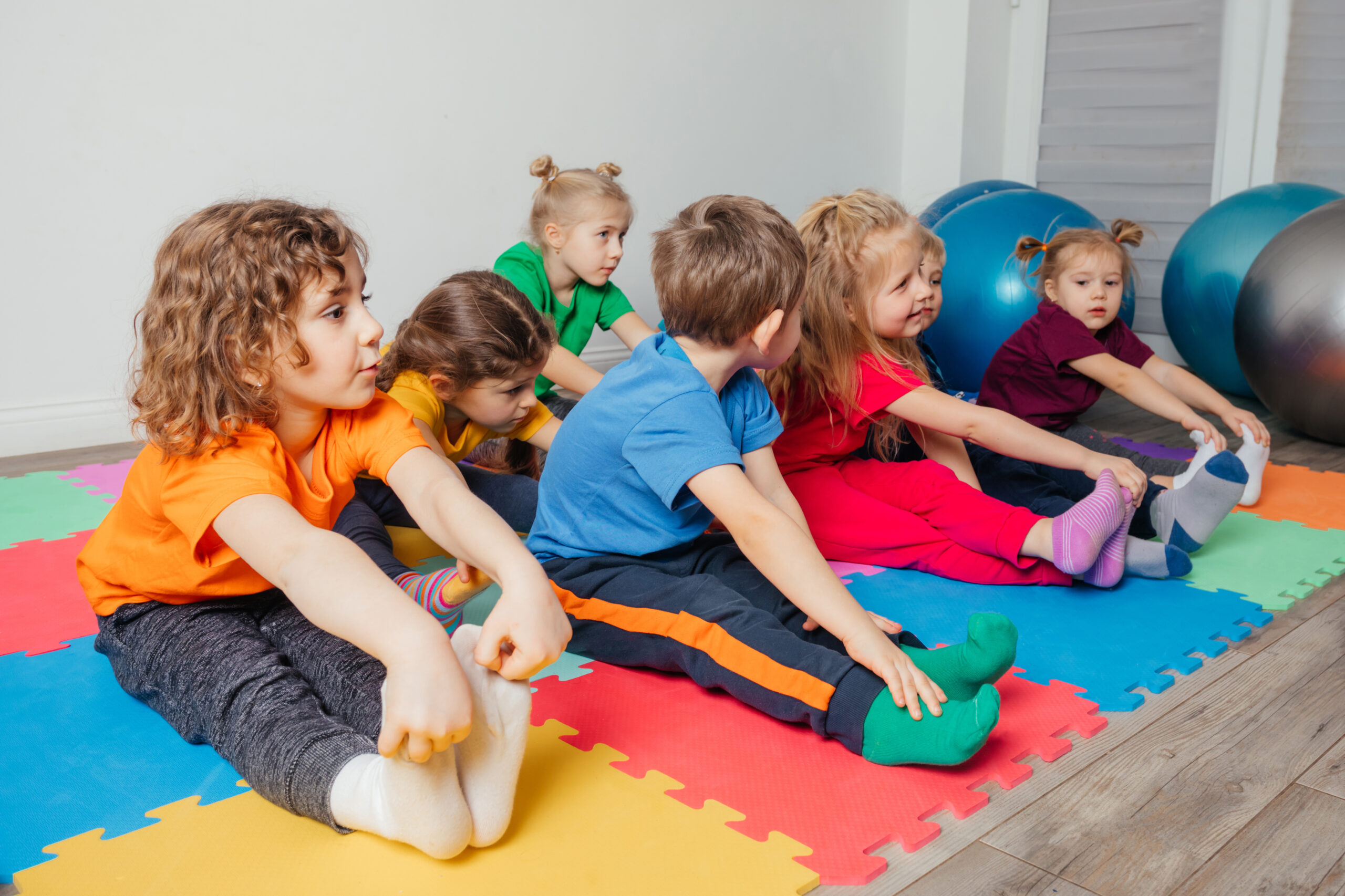 Adorable preschool kids in colorful clothes stretching legs sitting on a colourful mats. Yoga lesson for preschool children in modern and cosy gym.