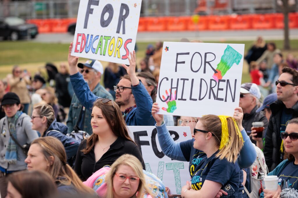 People stand at an outdoor rally holding signs that say "For Educators" and "For Children"